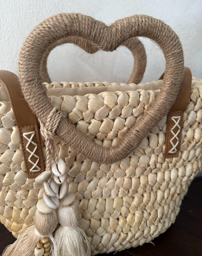 Capazo trenzado con asa en forma de corazón y detalles boho, perfecto para un estilo romántico y veraniego.